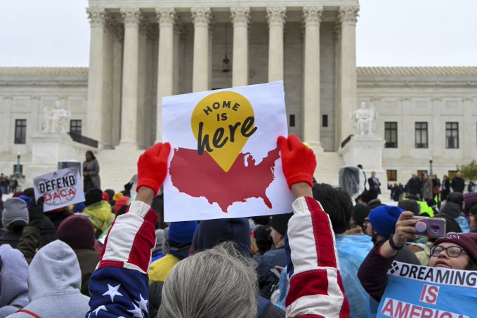 Protestors in front of the Supreme Court. One protestor in the center holds up a sign that says "home is here."